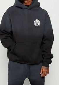 Sweat à capuche noir avec cordon de serrage, poche avant et logo circulaire sur la poitrine. Confectionné en tissu doux et texturé, avec une coupe décontractée.