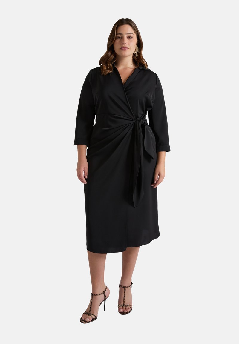 Robe noire à manches trois-quarts, avec une taille nouée et une texture lisse. Associée à des sandales à talons avec des lanières.