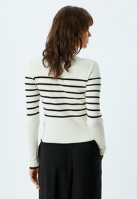 Maglione a coste di colore bianco sporco con strisce orizzontali nere. Vestibilità slim, maniche lunghe e scollatura rotonda. Indossato con pantaloni neri.