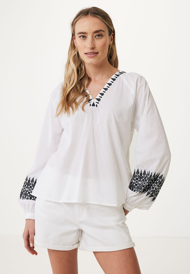 Blusa bianca a maniche lunghe con scollatura a V, caratterizzata da ricami neri lungo il collo e le maniche, realizzata in tessuto leggero. Abbinata a pantaloni corti bianchi.