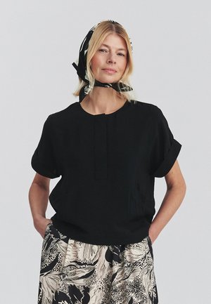 Blondynka ubrana w czarną bluzkę z krótkim rękawem, beżową i czarną wzorzystą spódnicę oraz czarną chustę z białymi wzorami zawiązaną na głowie.