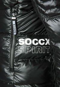 Giacca a vento nera con finitura testurizzata, chiusura con zip e audace testo bianco "SOCCX SPIRIT" sul pannello frontale.