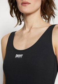 Zbliżenie kobiety w czarnej koszulce bez rękawów DKNY Sport z białym logo umieszczonym na środku klatki piersiowej.