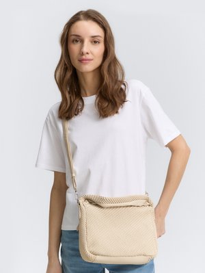 Sac à épaule tissé beige avec une surface texturée, une fermeture à rabat et une bandoulière ajustable, porté avec un t-shirt blanc et un jean.