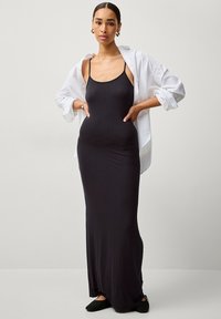 Schwarzes, figurbetontes Maxikleid aus glattem Stoff mit dünnen Trägern. Kombiniert mit einer weißen, lässigen Bluse mit aufgerollten Ärmeln.
