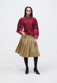 Röd blus med mönster, puffärmar och knappstängning fram, kombinerad med en khakifärgad veckad kjol. Modellen bär bruna strumpbyxor och vinröda klackskor.