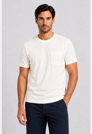 Mann mit kurzen dunklen Haaren, der ein weißes kurzärmeliges T-Shirt mit Tasche und schwarze Shorts trägt, steht vor einem schlichten grauen Hintergrund.