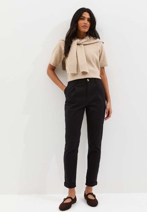 Kobieta o długich ciemnych włosach, ubrana w beżowy sweter z krótkim rękawem i pasującym szalikiem, czarne spodnie z wysokim stanem oraz brązowe baleriny, stojąca przy ścianie.