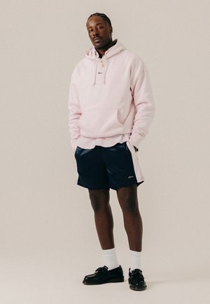Homme avec les cheveux tressés portant un sweat à capuche rose clair, un short bleu marine, des chaussettes blanches et des mocassins noirs, debout les mains dans les poches sur un fond neutre.