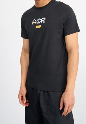 Mann trägt schwarzes Kurzarm-T-Shirt mit mehrfarbigem "A C R" und gelbem Nike-Logo auf der Brust, kombiniert mit schwarzen Hosen.