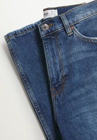 Jean en denim bleu foncé légèrement délavé, avec des coutures orange, un bouton avant et une étiquette indiquant les tailles pour différentes régions.