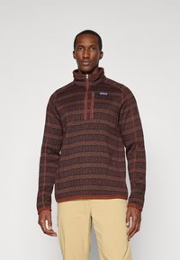 Fleece pullover with a high collar, featuring a patterned design in dark red, green, and brown. Includes a chest zipper and logo detail.
