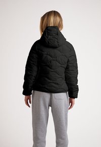 Giubbotto a vento nero con cappuccio, caratterizzato da una texture trapuntata e polsini elasticizzati, indossato sopra pantaloni tuta grigi, visto da dietro.