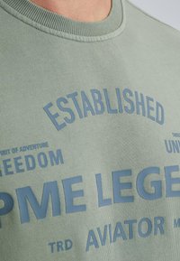 Zelená mikina s rebrovaným výstrihom, s modrým potlačeným textom vrátane "ESTABLISHED" a "PME LEGEND" na prednej strane.