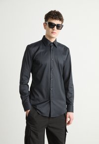 Camisa negra de botón con una textura suave, mangas largas y un collar puntiagudo. Combinada con gafas de sol negras, sobre un fondo neutro.