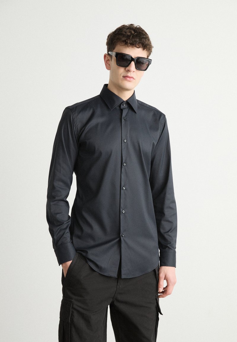 Camisa negra de botón con una textura suave, mangas largas y un collar puntiagudo. Combinada con gafas de sol negras, sobre un fondo neutro.