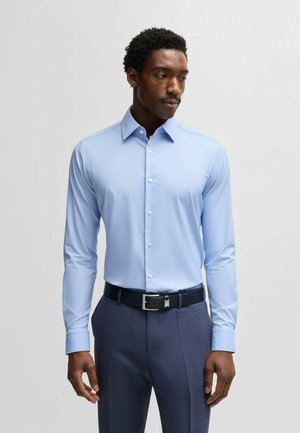 Chemise bleu clair à manches longues avec col boutonné, présentant une texture lisse et un design ajusté, associée à un pantalon foncé et une ceinture.