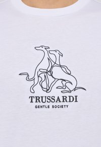 Biała koszulka z czarnym rysunkiem dwóch psów oraz tekstem "TRUSSARDI GENTLE SOCIETY" poniżej w pogrubionej czarnej czcionce. Miękka w dotyku.