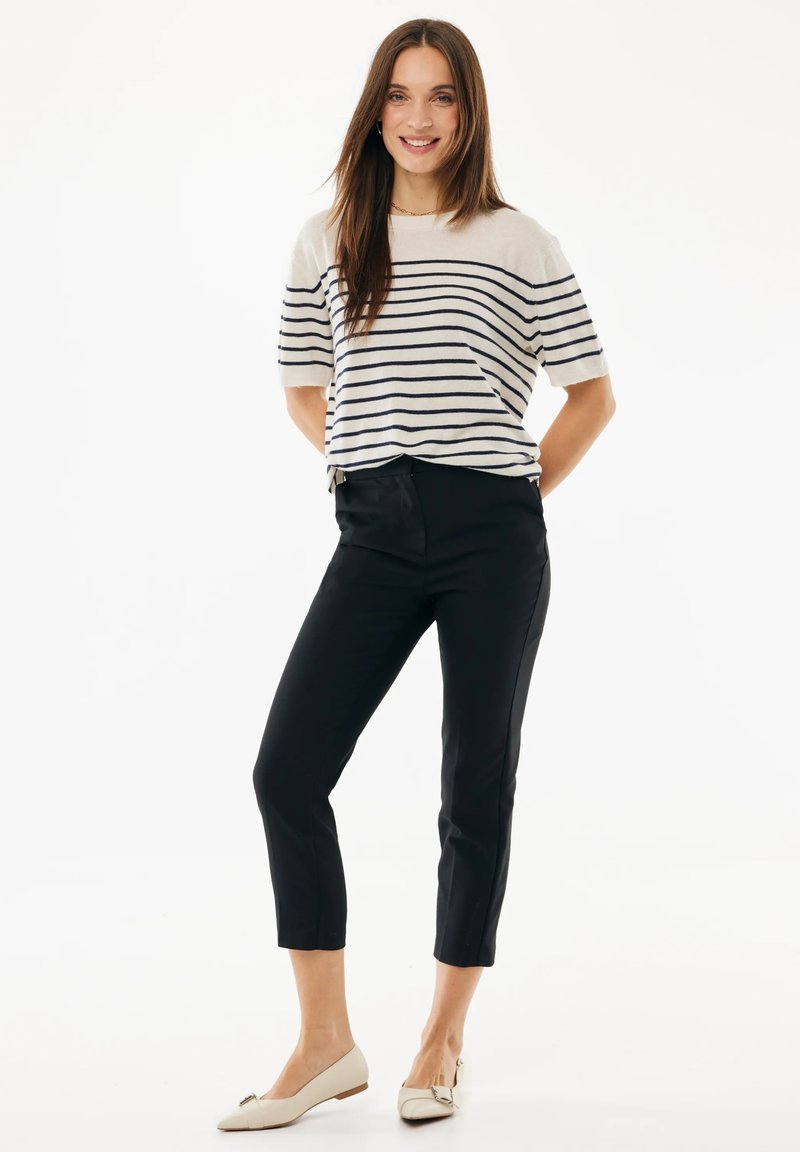 Mujer de pie sonriendo, vestida con un suéter crema de manga corta con rayas azul marino, pantalones negros recortados y zapatos planos beige con punta.