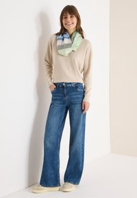 Beige trui, wijde blauwe jeans en een sjaal met een patroon in groen, blauw en wit. Casual schoenen maken de outfit compleet.