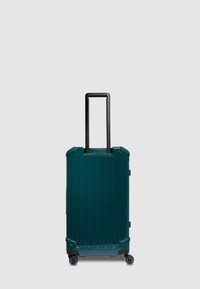 Maleta de viaje rígida en verde azulado oscuro, con ranuras verticales, asa retráctil negra y cuatro ruedas giratorias para facilitar la movilidad.