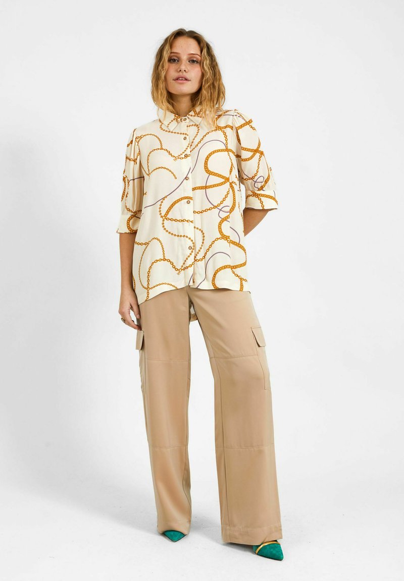 Femme portant un chemisier crème à motif chaîne orange, un pantalon cargo beige à jambes larges, et des chaussures vertes pointues, debout devant un fond blanc.