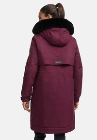 BURGUNDAS ziemas parka ar lielu melnu mākslīgās kažokādas apkakli, kapuci un zīmola birku. Tā ietver garas piedurknes un elastīgas aproces.