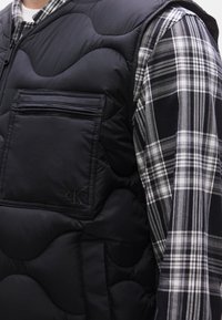 Μαύρο αμάνικο μπουφάν με κουκούλα και ραμμένο σχέδιο, με τσέπη στο στήθος με φερμουάρ. Φορεμένο πάνω από ένα καρό πουκάμισο σε μαύρο και άσπρο. Υφασμάτινη υφή.