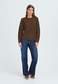 Brauner gestrickter Pullover mit einem strukturierten Muster, gerippten Bündchen, getragen mit blauen Jeans und braunen Sneakers, stehend vor einem hellen Hintergrund.