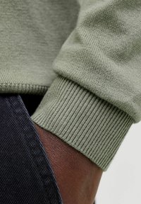 Poignet de pull vert tricoté avec texture côtelée, adjacent à un tissu foncé, mettant en valeur le tissage doux et les couleurs contrastantes des matériaux.