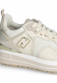 Beige sportlicher Schuh aus Mesh und Leder, mit einer strukturierten Oberfläche, goldener Logo-Akzent und einer dicken weißen Sohle mit profilierter Unterseite.