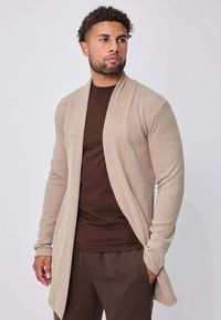 Homme portant un cardigan beige ouvert sur une chemise et un pantalon marron foncé, debout devant un fond uni gris clair, regardant légèrement vers la gauche.