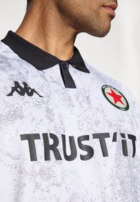 Gros plan sur une personne portant un polo texturé blanc et gris avec un col noir, arborant le texte "TRUST'IT", un logo en étoile rouge et le symbole Kappa.