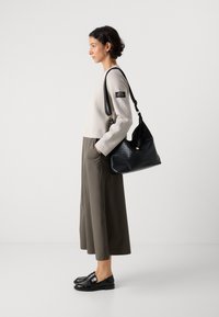 Mujer con una blusa de manga larga beige, pantalones anchos verdes oliva, mocasines negros, llevando un bolso de hombro de mimbre negro, de pie de lado sobre un fondo blanco.