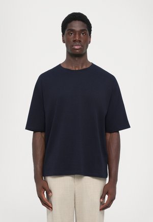 Tumed sinine lühikeste varrukatega T-särk lihtsa disainiga ümmarguse kaelusega, valmistatud siledast kangast, mida on kombineeritud heledate beežide pükstega.