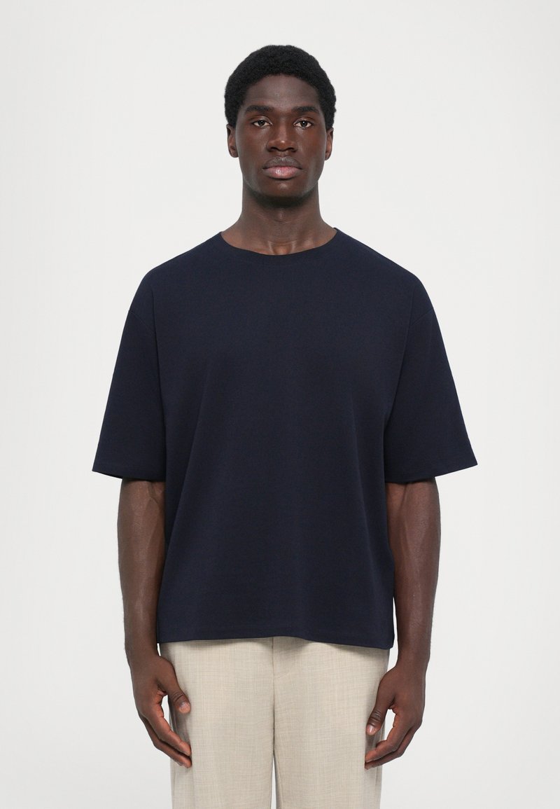 Σκούρο μπλε κοντομάνικο μπλουζάκι σε απλό σχέδιο με στρογγυλή λαιμόκοψη, φτιαγμένο από λείο ύφασμα, συνδυασμένο με ανοιχτό μπεζ παντελόνι.