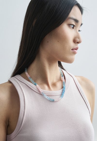 Κολιέ με χάντρες που διαθέτει παστέλ μπλε, ροζ και πράσινες πέτρες, τακτοποιημένες σε ένα απαλό, κεκλιμένο σχέδιο, φορεμένο πάνω από ένα ανοιχτό ροζ ριμπά μπλουζάκι.