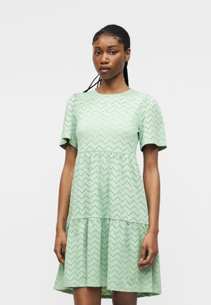 Młoda kobieta ubrana w miętowo zieloną sukienkę z krótkim rękawem i zygzakowatym wzorem oczek, stojąca na jasnym, jednolitym tle.