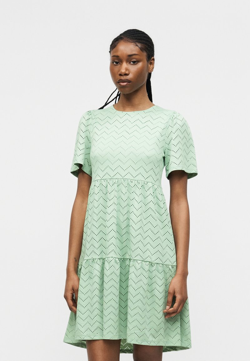 Νεαρή γυναίκα που φορά ένα τιρκουάζ κοντομάνικο φόρεμα με ζιγκ-ζαγκ σχέδιο με τρύπες, στέκεται σε απλό ανοιχτό φόντο.