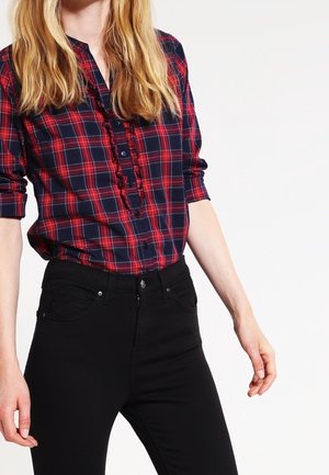 Femme portant une chemise à carreaux rouge et marine à manches longues rentrée dans un pantalon noir taille haute, debout sur fond blanc.