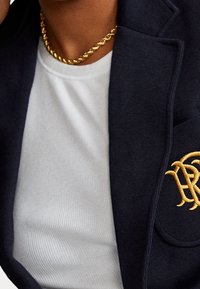 Blazer azul marino con bordados dorados en el bolsillo, llevado sobre una camiseta blanca de canalé. Un collar de cadena dorada añade un toque texturizado.