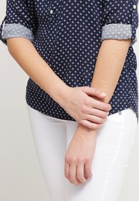 Kvinna iklädd en marinblå mönstrad skjorta och vita byxor, med händerna försiktigt knäppta runt vänster handled i midjehöjd.