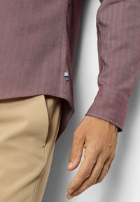 Bordeauxfarbenes Hemd mit Fischgrätmuster und langen Ärmeln, mit einem blauen, weißen und grauen Akzentetikett am Saum. Kombiniert mit beigen Hosen.