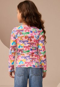 Langarmhemd aus geripptem Stoff mit einem mehrfarbigen Schleifendruck auf einem rosa Hintergrund, kombiniert mit Jeans.