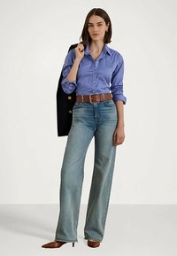 Γυναίκα που φοράει ένα μπλε πουκάμισο με κουμπιά, τσέπεται σε ψηλή μέση φαρδεί παντελόνια με καφέ ζώνη και τακούνια, κρατώντας μια μαύρη ζακέτα στον ώμο.