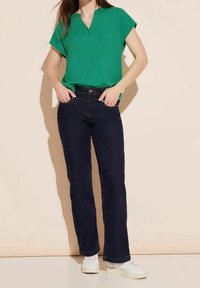 Donna che indossa una maglietta verde a maniche corte con scollatura a V, jeans dritti blu scuro e sneakers bianche, in piedi con le mani nelle tasche.