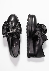 Scarpe in pelle nera con un grande fiocco testurizzato sulla parte anteriore, su una suola spessa e rigata e una finitura lucida.