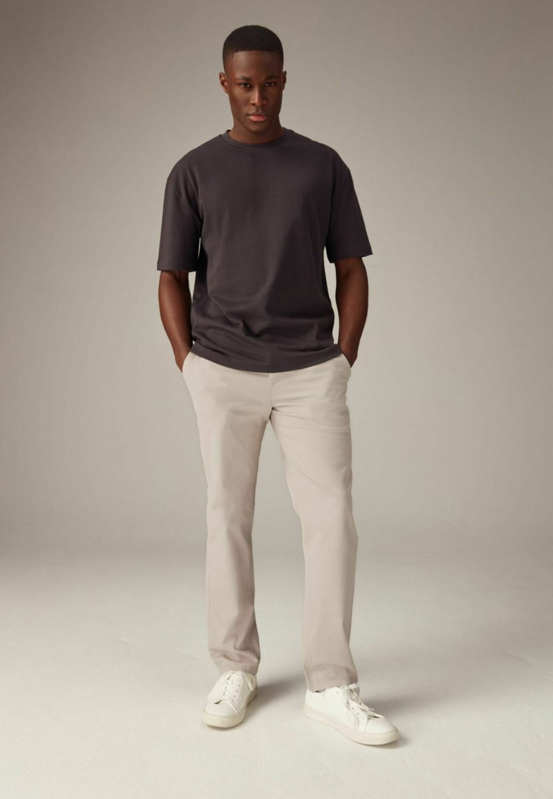 T-shirt grigio scuro a maniche corte con una vestibilità rilassata, abbinato a pantaloni beige chiaro e sneaker bianche, posato su uno sfondo neutro.