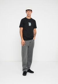 Black t-shirt with graphic print in blue, paired with grey baggy jeans. Model wearing black sneakers and a white cap stands against a plain background.
