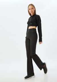 Zwarte cropped hoodie met trekkoorden, gecombineerd met hooggewaaid zwarte flare broeken. Model draagt zwarte sneakers met grijze zolen.